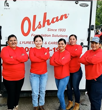 Five female members of an all-women foundation repair crew standing in front of an Olshan Foundation Solutions service truck, wearing matching red long-sleeved shirts and smiling.