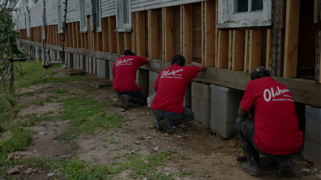 Three Olshan Foundation Solutions technicians in red shirts working on a foundation repair, adjusting concrete blocks and wooden beams to level a hotel