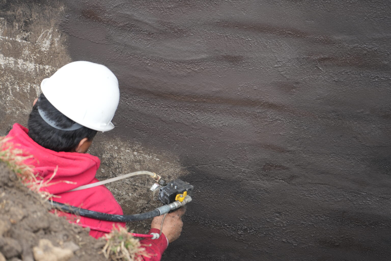 Olshan Certified Structural Technician spraying rubberized WetSuit coating onto a basement wall from the trench